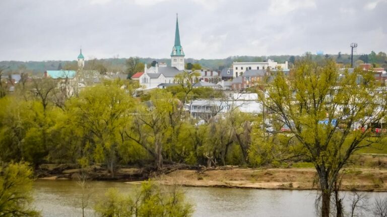 Panoramic view of a small town with church steeples, green trees, and a river in the foreground.