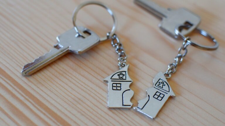 Two silver house-shaped keychains with keys, placed on a light wooden surface.