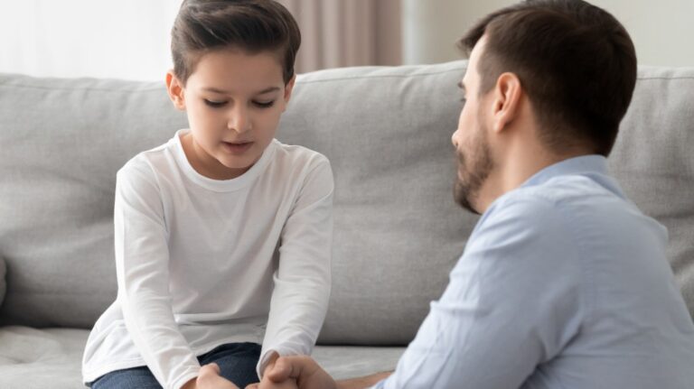 Young boy in white shirt sitting on gray couch, holding hands with adult male.