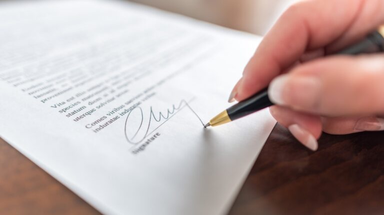 Close-up of a hand signing a document with a black and gold pen on a wooden surface.