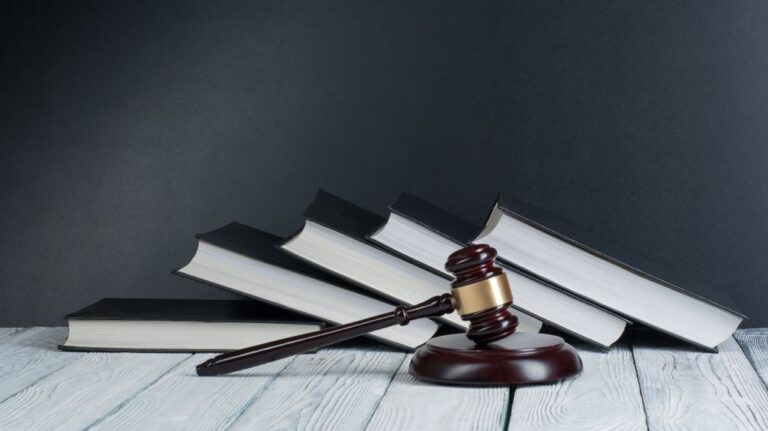 Wooden judge's gavel resting in front of a stack of black hardcover legal books on a white wooden surface.