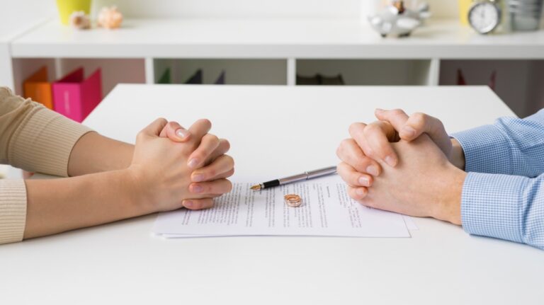 Two people with clasped hands sit across a white table with a contract, pen, and two wedding rings.