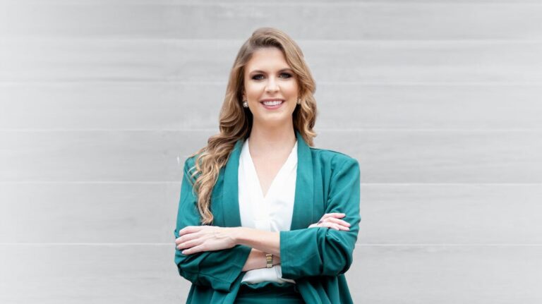 Professional woman with blonde hair wearing a teal blazer and white blouse, smiling with arms crossed.