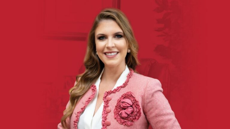 Professional portrait of a smiling woman with long hair wearing a pink blazer with floral details against a red background.