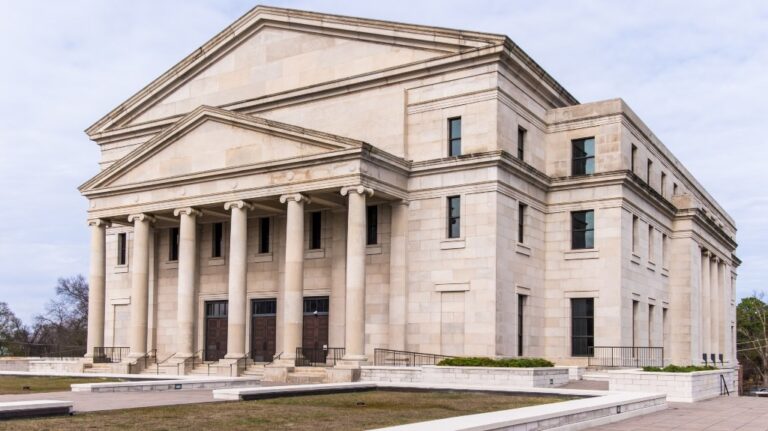 Large neoclassical stone building with tall columns and multiple windows under a cloudy sky.