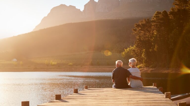 Elderly couple sitting on wooden dock by lake at sunset with mountains and trees.