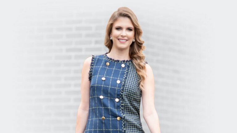 Woman with long blonde hair wearing a sleeveless blue checkered dress with gold buttons.