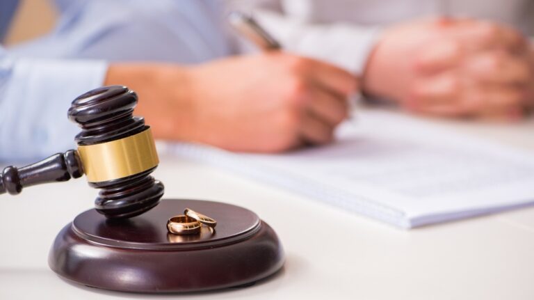 Close-up of a wooden judge's gavel and two gold wedding rings on a desk with a person writing in the background.