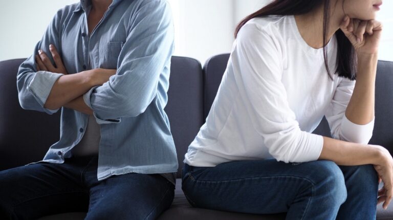 Two individuals sitting back-to-back on a dark gray sofa, both appearing upset and avoiding eye contact.
