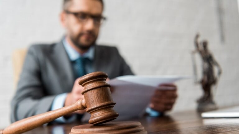 Wooden judge's gavel in foreground with blurred man in suit reviewing documents behind.