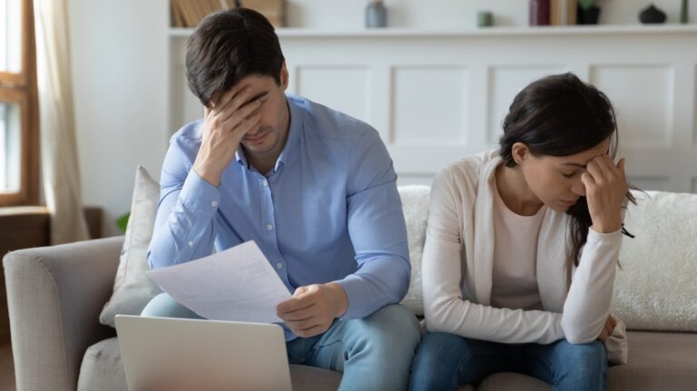 Man and woman sitting on sofa, both holding their heads in distress, with documents and laptop.