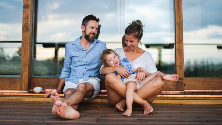 Family of three sitting on wooden deck in front of glass doors, smiling and relaxed.