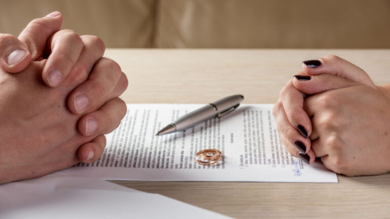 Two pairs of hands clasped on a table beside a signed document, a pen, and two wedding rings.