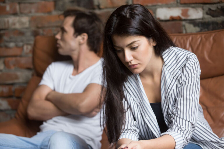 A young woman in a striped jacket looks down thoughtfully while a man in a white shirt sits with arms crossed, both appearing upset.