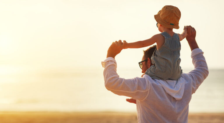 Man wearing glasses carrying child in striped overalls and brown hat on shoulders at beach during sunset.