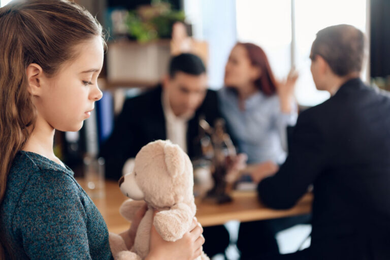 Young girl holding a teddy bear in the foreground with three adults in a discussion in the background.