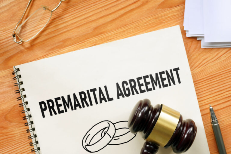 Spiral-bound premarital agreement document with wedding rings illustration, wooden gavel, eyeglasses, and pen on desk.