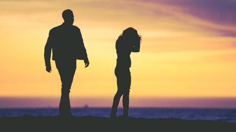 Silhouettes of a man and woman standing near the ocean at sunset with an orange sky.