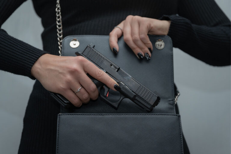 Close-up of a person with black nail polish holding a black pistol inside a black handbag.