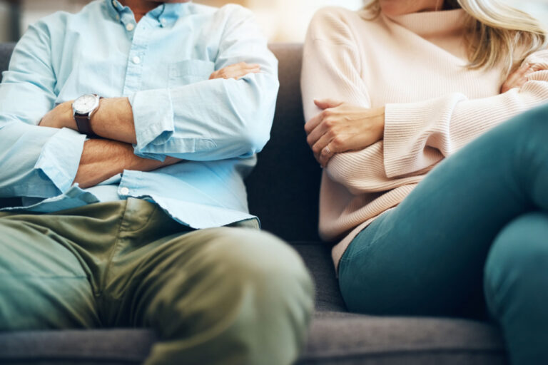 Two people sitting on a gray couch with arms crossed, wearing casual clothing.