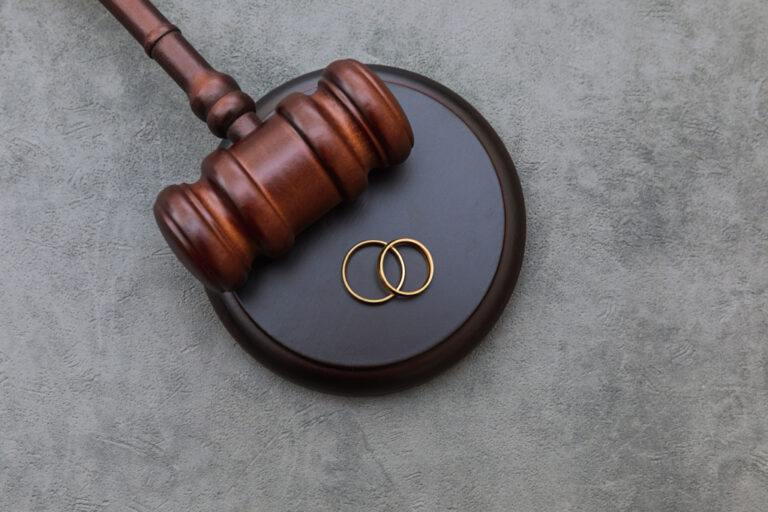 Wooden judge's gavel resting on base with two intertwined gold wedding rings on gray background.