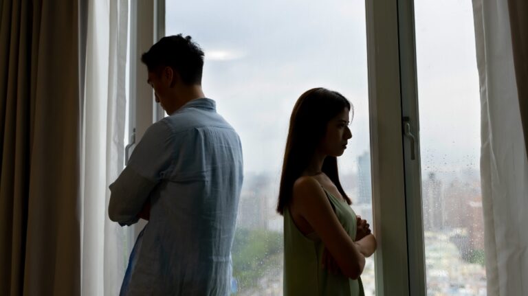 Silhouetted man and woman standing back-to-back by rain-speckled window, appearing upset.