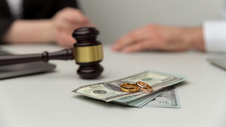 Two gold wedding rings on a stack of US dollar bills with a judge's gavel and blurred hands in the background.