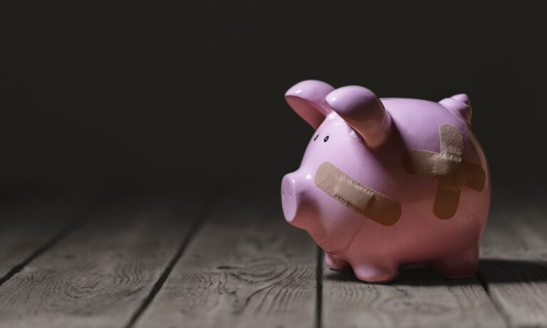 Pink ceramic piggy bank with adhesive bandages on wooden floor against dark background.