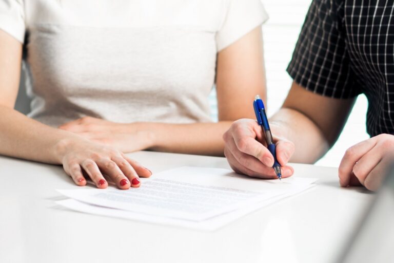 Two people sitting at a white table, one signing a document with a blue pen.