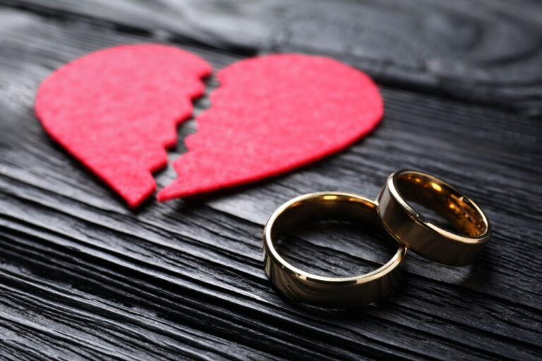 Two gold wedding rings placed beside a broken red heart on dark wooden surface.