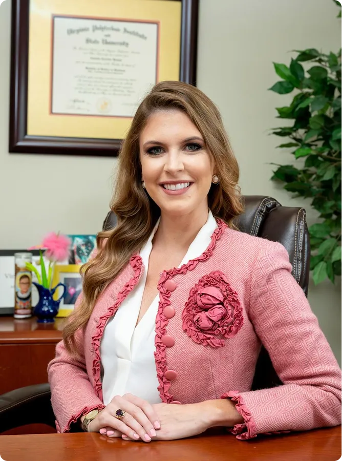 Professional woman with long wavy hair wearing pink jacket, seated at desk with diploma and plant in background.