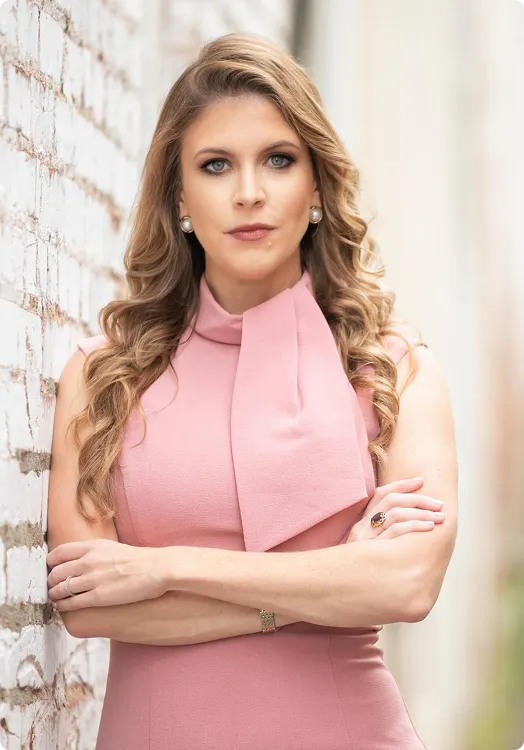 Professional woman with long blonde hair wearing a pink sleeveless dress, standing against a white brick wall with arms crossed.