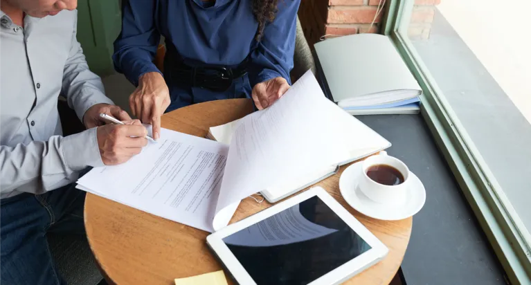 Two people reviewing and signing documents at a round wooden table with a tablet and coffee cup.