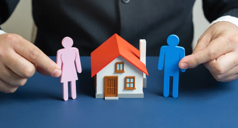 Two hands holding pink female and blue male figurines next to a small white house with a red roof on a blue surface.