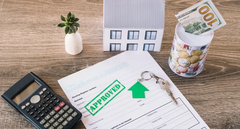 Calculator, miniature house, approved document with key and green arrow, and money jar on wooden table.