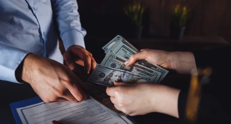 Two people exchanging US dollar bills over a document on a clipboard.