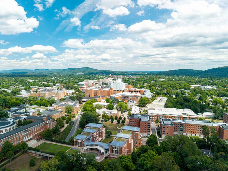 Aerial view of a university campus with multiple brick buildings, green trees, and hills under a partly cloudy sky.