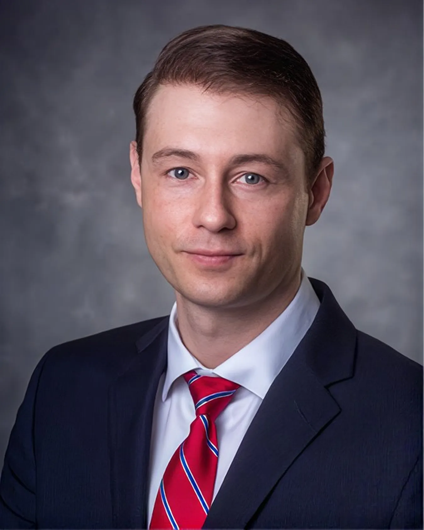Professional headshot of a man in a dark suit, white shirt, and red striped tie.
